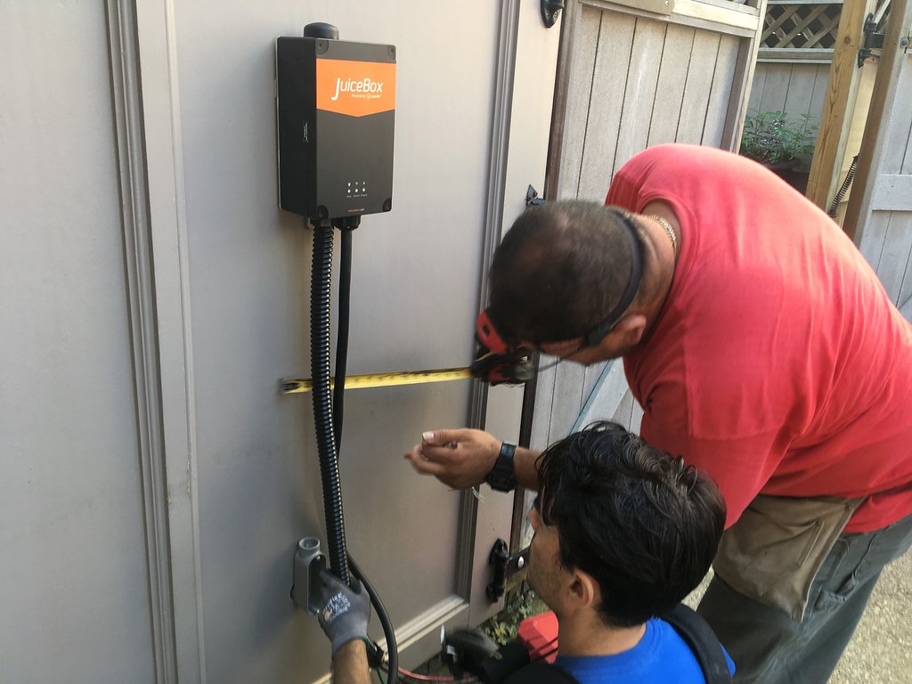 Two technicians installing a JuiceBox EV charger mounted on a gray garage wall, measuring placement with a tape measure.