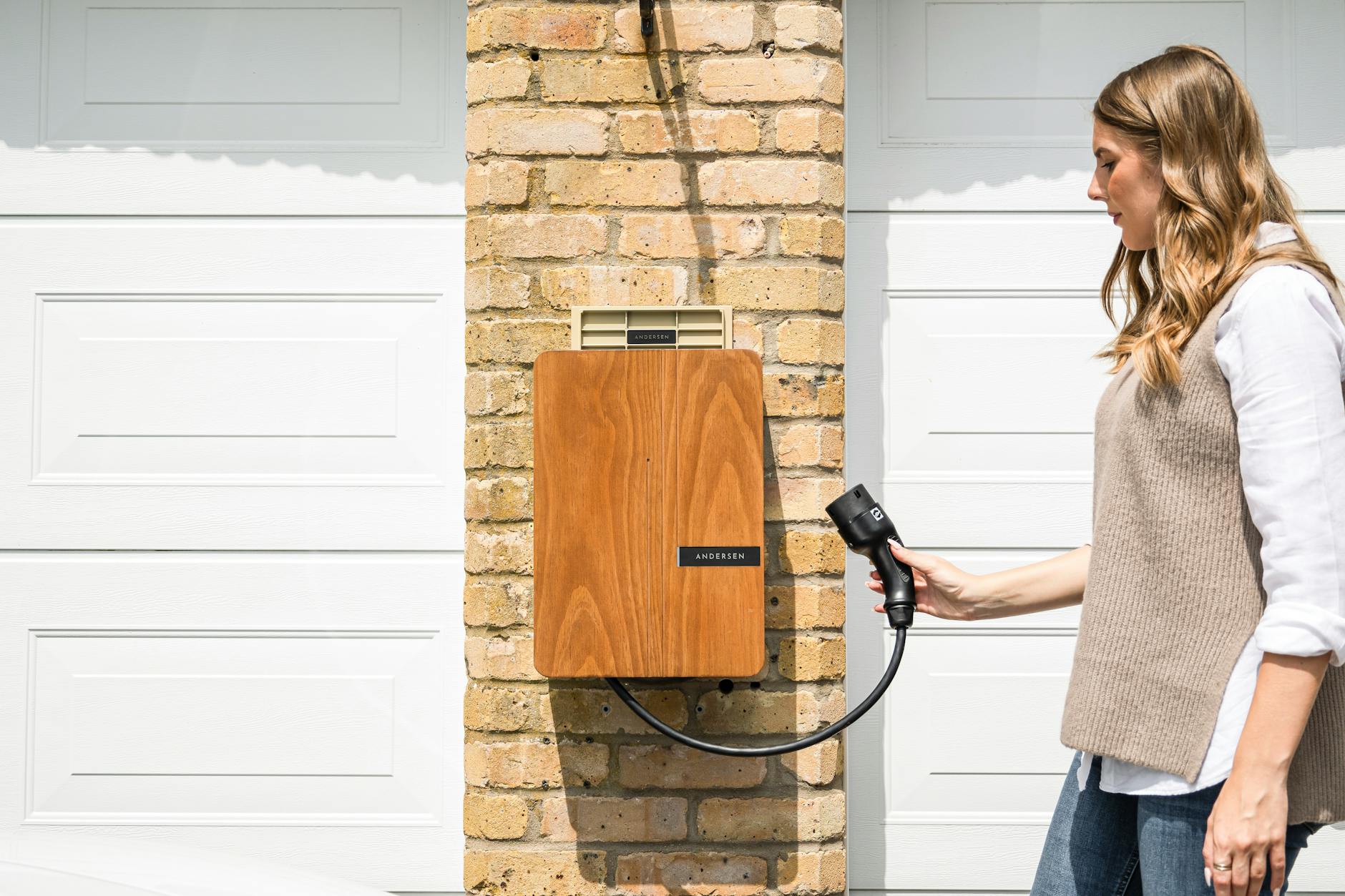 Woman holding charging cable connected to wood-paneled Andersen wallbox EV charger mounted on brick garage wall