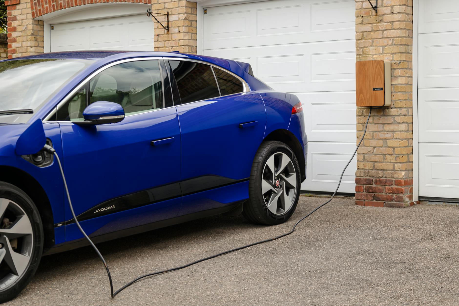 Orange wall-mounted EV charger installed on brick wall next to garage door, charging cable connected to blue Jaguar electric sedan.