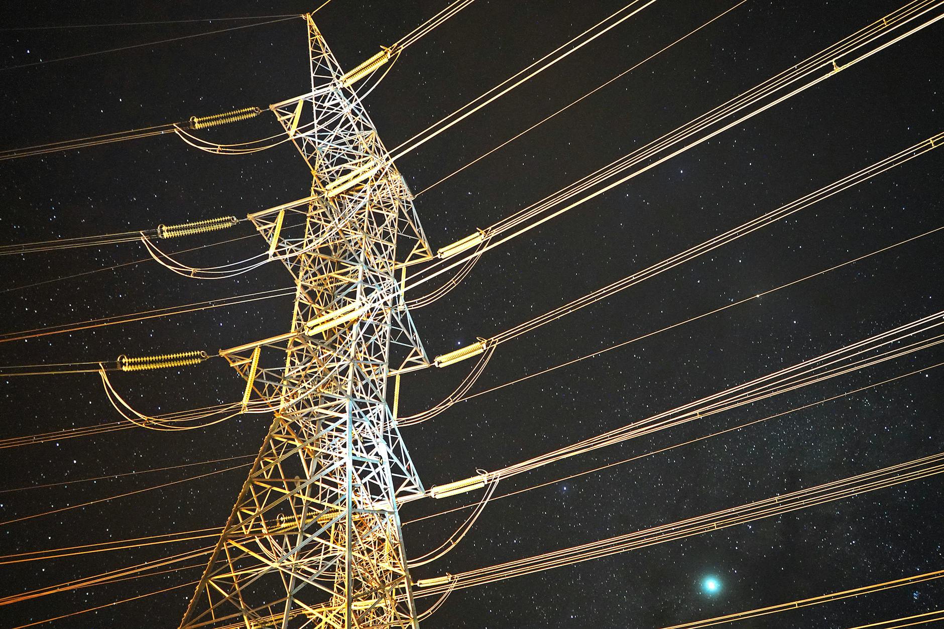 High voltage transmission tower with multiple power lines radiating outward, photographed from below against a dark sky.