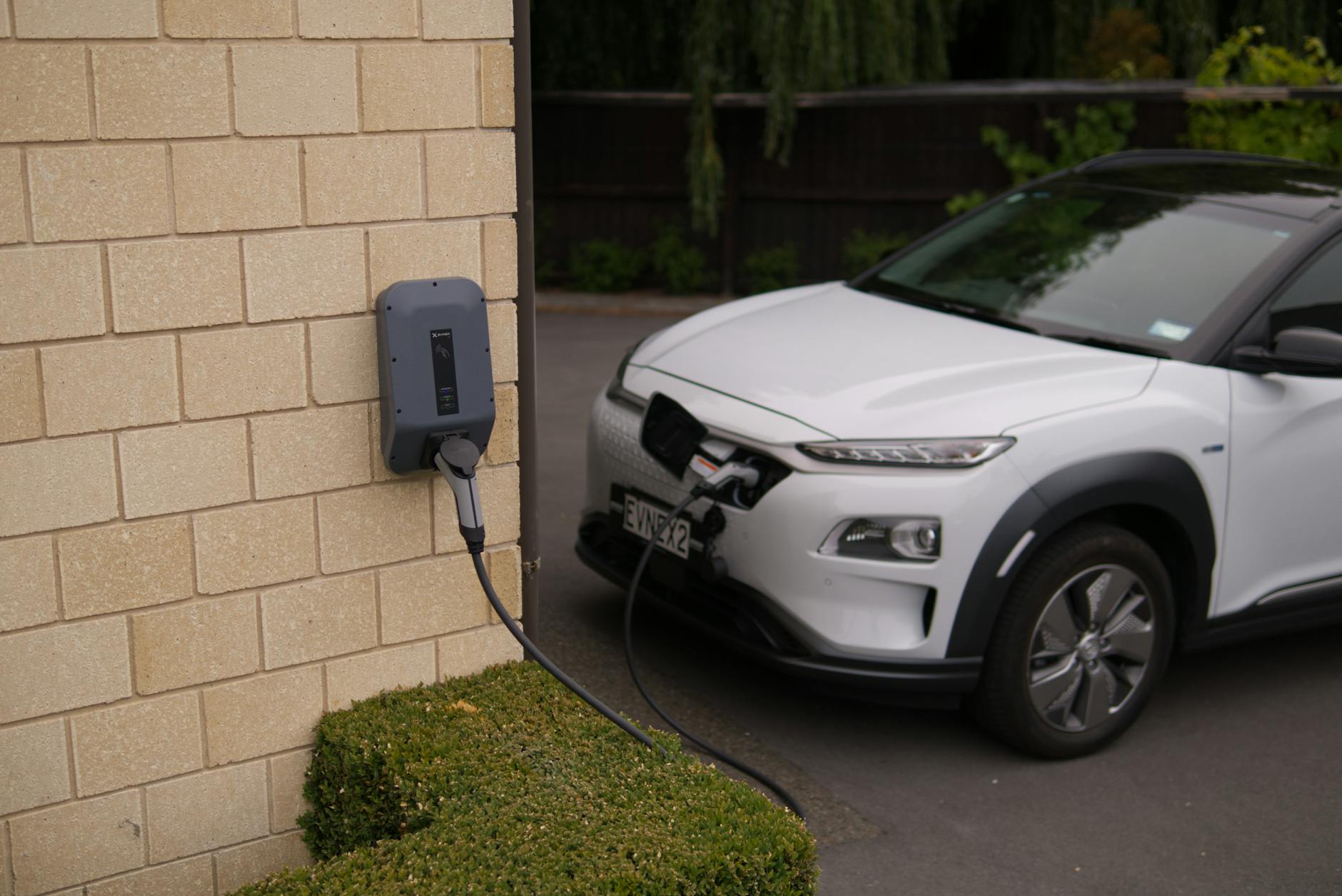 Wall-mounted EV charger installed on brick exterior with charging cable connected to white electric vehicle in parking bay