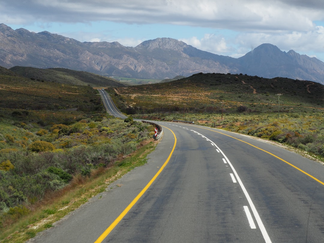 Open highway with yellow center lines stretching through semi-arid landscape with dramatic mountain ranges in the background