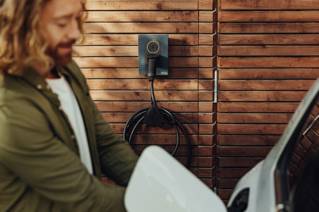 Wall-mounted EV charger with coiled black cable on wood-paneled exterior wall, person reaching for charging connector