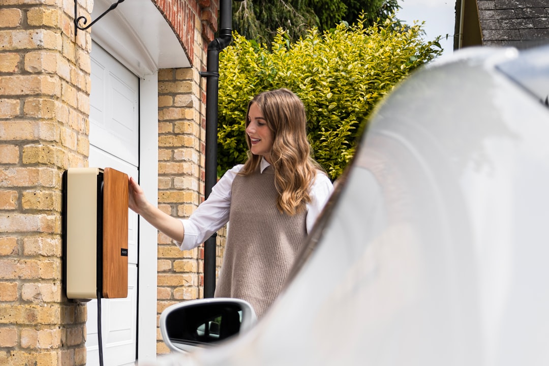 Woman using a wall-mounted EV charger with wooden accent panel on brick garage wall, white electric vehicle plugged in nearby