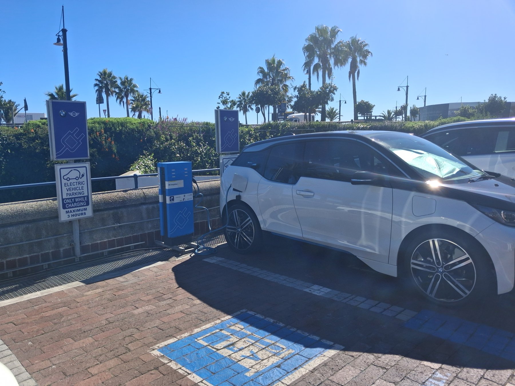 A white BMW i3 plugged into a public EV charger at the V&A Waterfront in Cape Town