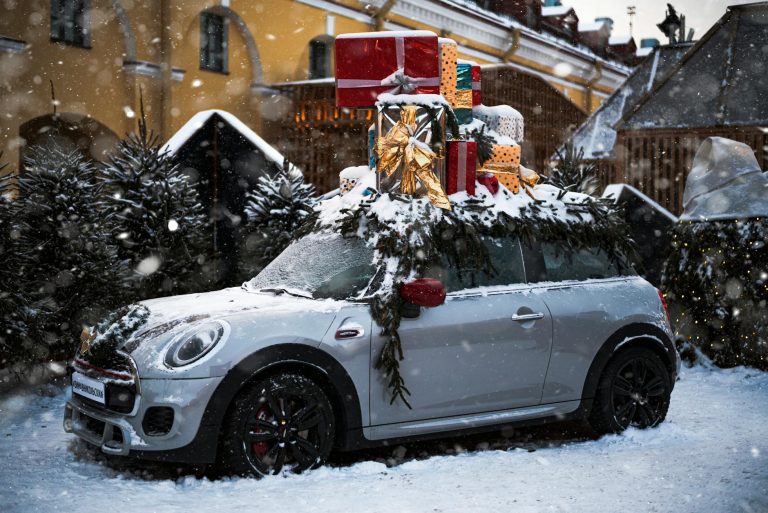 A Mini Cooper loaded with Christmas gifts parked in snowy outdoors decorated with evergreen branches.