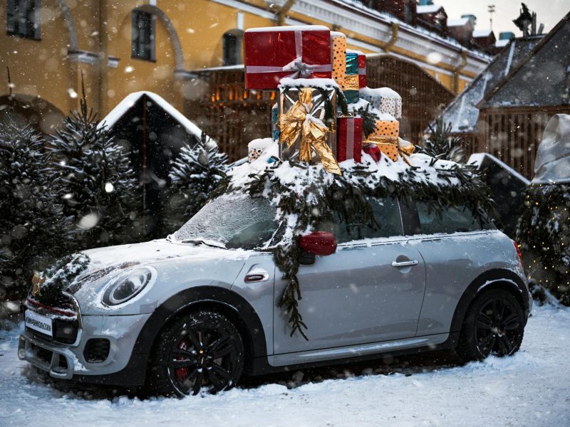 A Mini Cooper loaded with Christmas gifts parked in snowy outdoors decorated with evergreen branches.