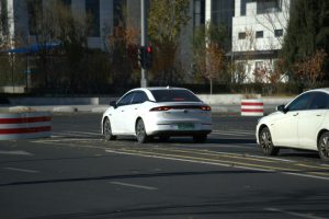 Two white cars driving on a city road with autumn trees in the background.