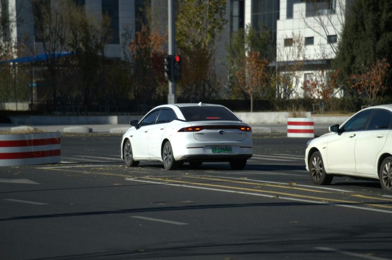 Two white cars driving on a city road with autumn trees in the background.