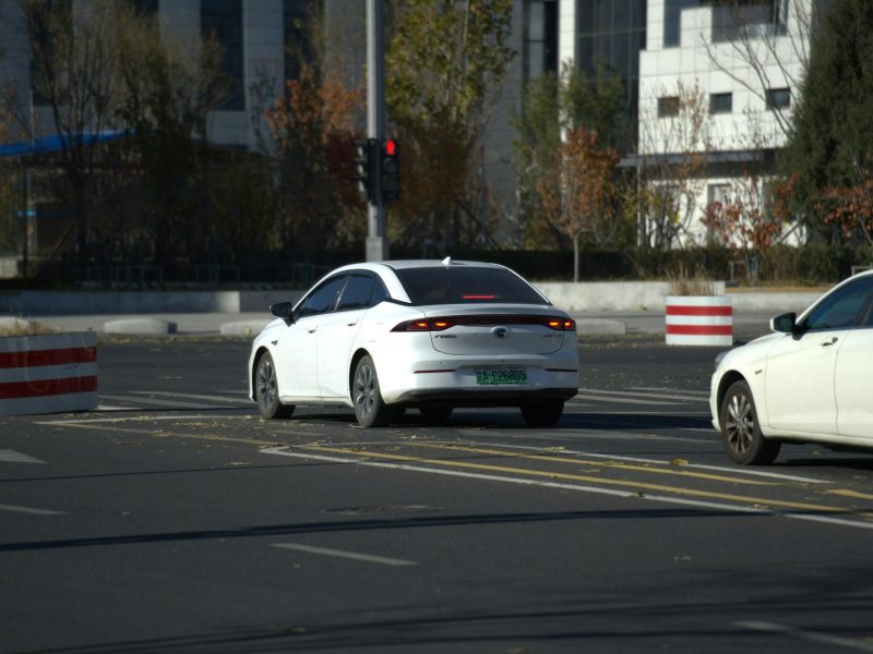 Two white cars driving on a city road with autumn trees in the background.
