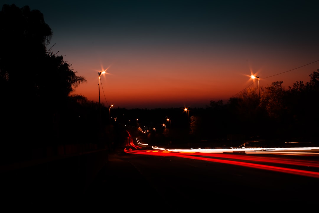 Suburban street at dusk with car light trails and street lamps illuminating a residential neighborhood under an orange-pink sky