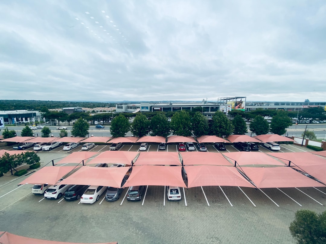 Aerial view of covered parking lot with red canopies sheltering rows of vehicles, modern commercial buildings visible in background.