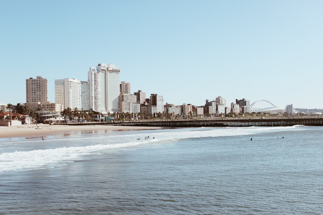Durban beachfront with skyline of high-rise buildings, sandy beach, waves, and the iconic Moses Mabhida Stadium arch visible on horizon