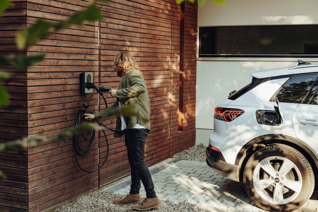Person plugging charging cable from wall-mounted EV charger into white electric vehicle parked in residential driveway