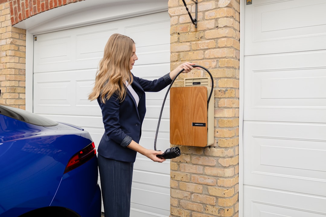 Woman holding charging cable from wood-finish wall-box EV charger mounted on brick garage wall beside parked electric vehicle