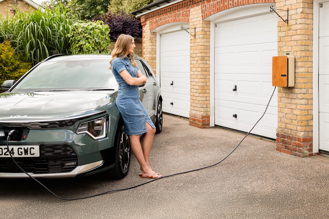 Electric vehicle plugged into orange wall-mounted charging unit outside a residential garage with person standing nearby