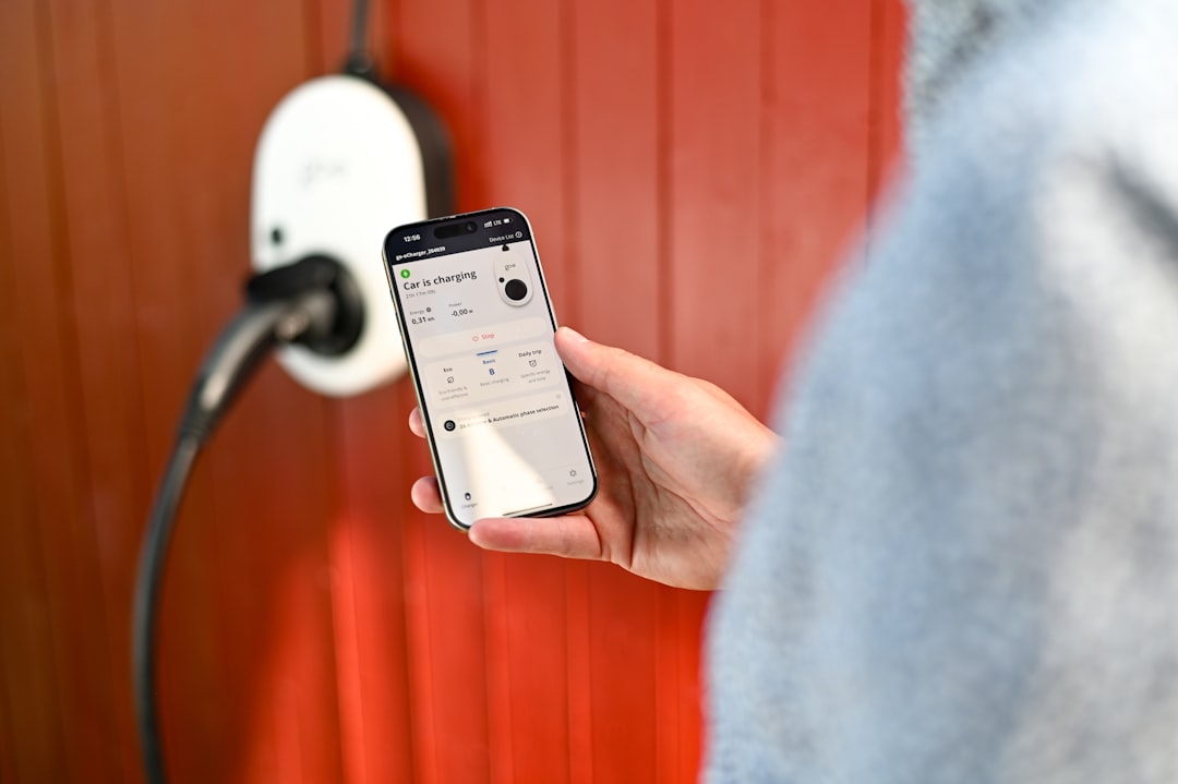 Wall-mounted white EV charger unit on orange wall with person holding smartphone showing active charging session details