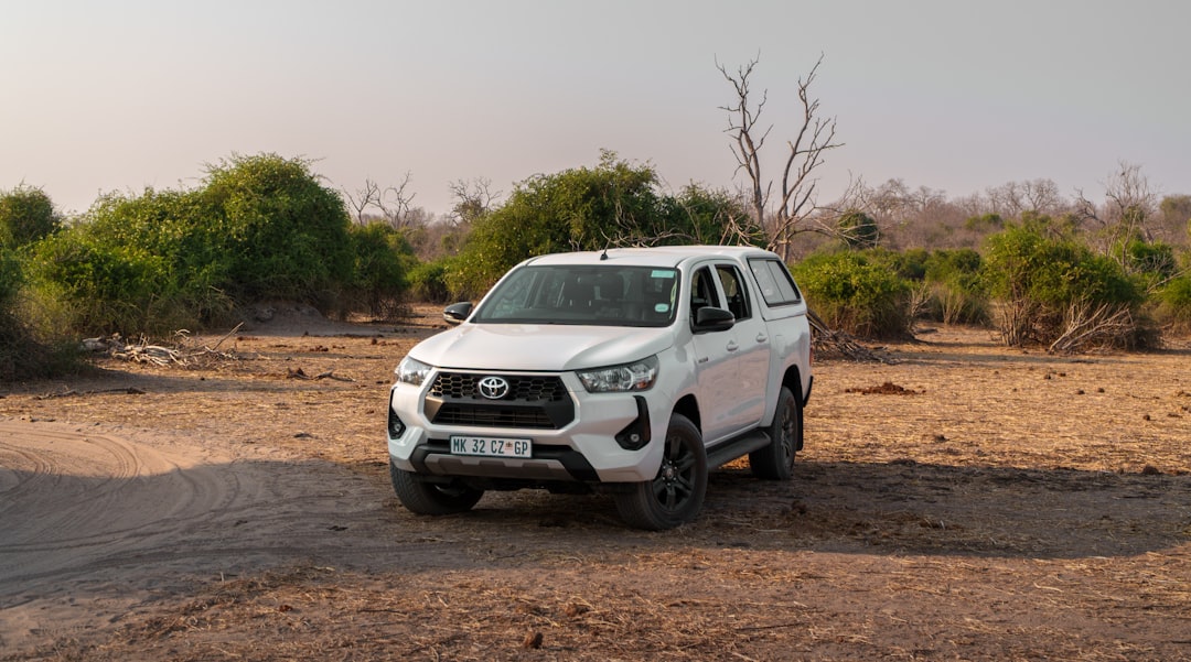 White Toyota Hilux double-cab pickup truck on sandy terrain with sparse vegetation in background.