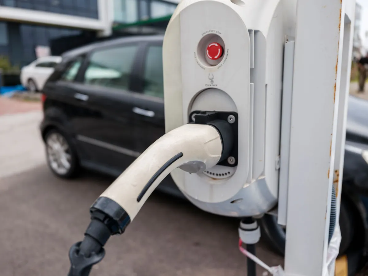 EV charging station with white connector mounted on post in parking lot, with parked vehicles in background
