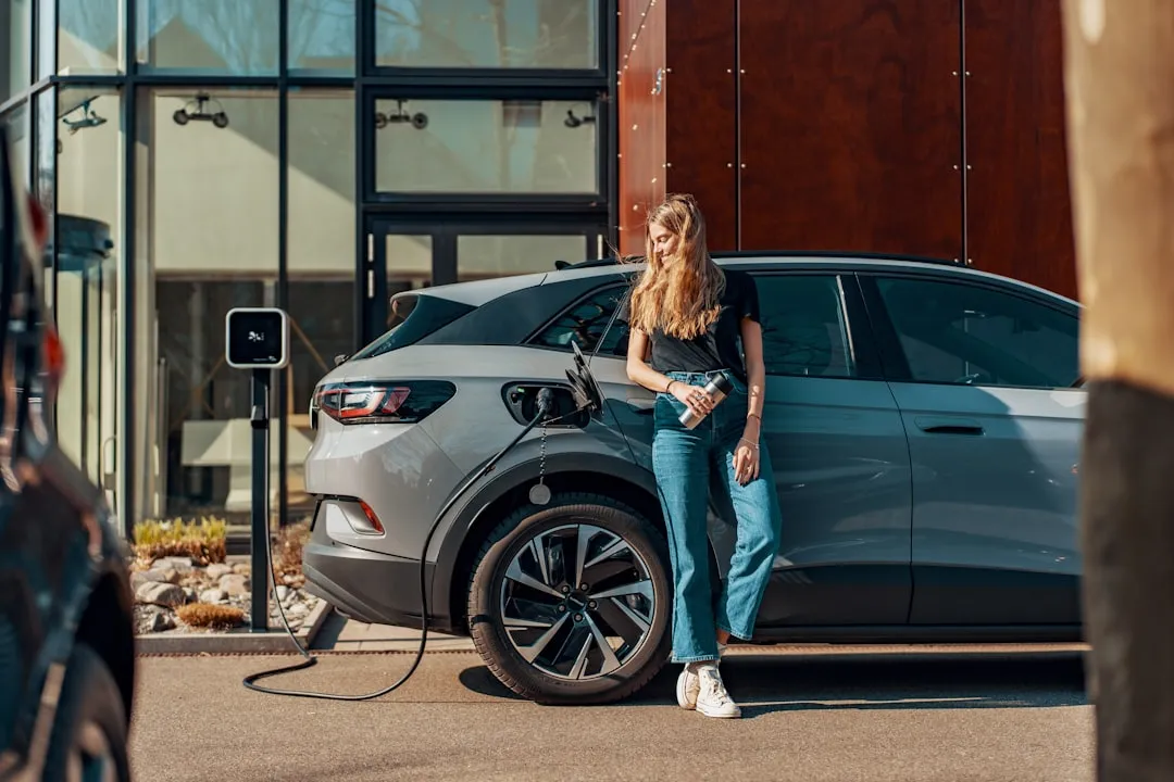 Woman standing beside white electric car connected to public charging station in urban setting
