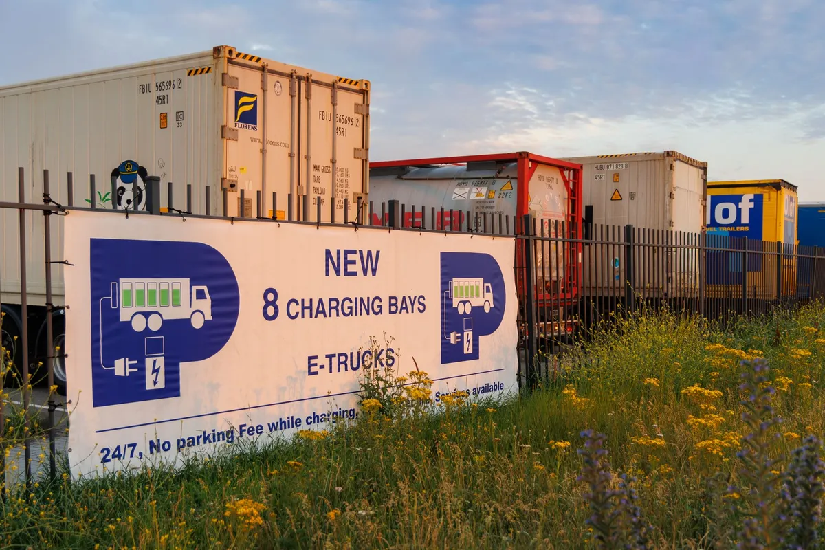 Modern EV charging station with 8 bays for electric trucks, blue and white signage, industrial facility with shipping containers and fencing, South African landscape