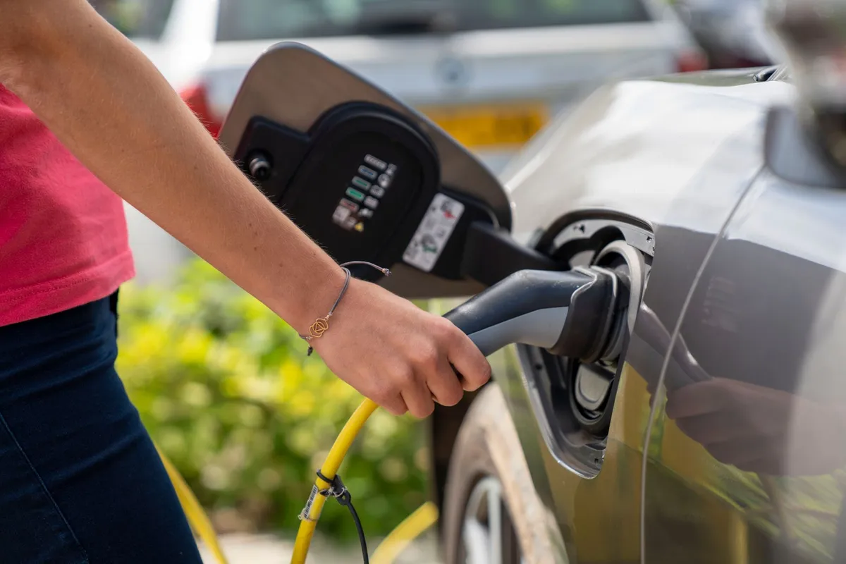 Person in pink shirt inserting yellow EV charging connector into vehicle port at retail charging station with display interface visible