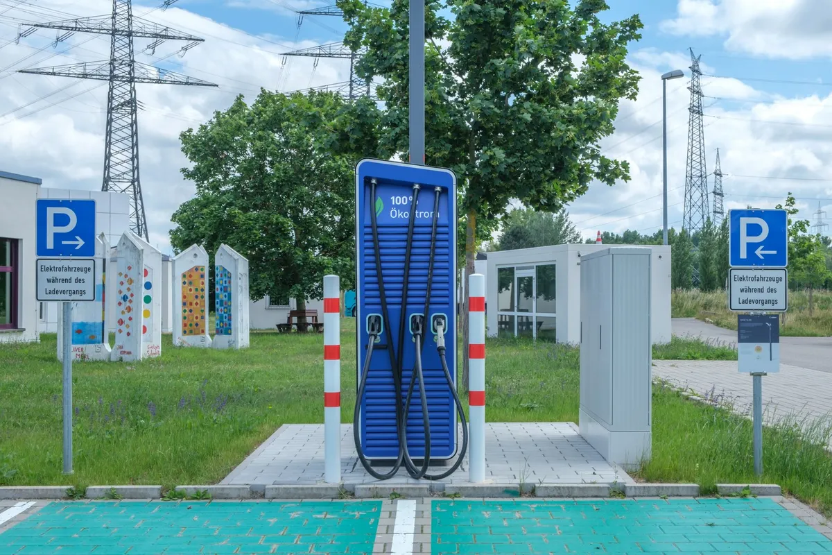 Blue dual-pedestal EV fast charger with two charging cables and connectors, installed on concrete pad with red-white bollards, parking signs, and electrical infrastructure visible.
