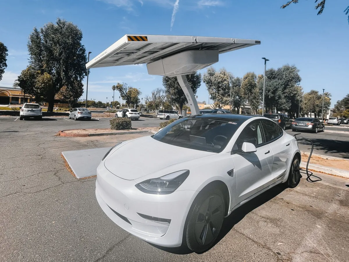 White Tesla Model 3 charging at a solar-canopy EV charging station in a commercial parking lot with trees and vehicles in background