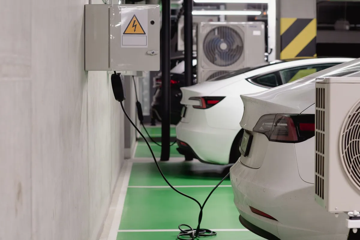 Electric vehicles charging in depot with wall-mounted electrical panel and black charging cables on green floor markings
