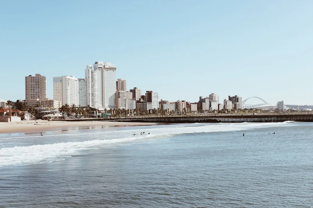 Durban beachfront with skyline of high-rise buildings, sandy beach, waves, and the iconic Moses Mabhida Stadium arch visible on horizon