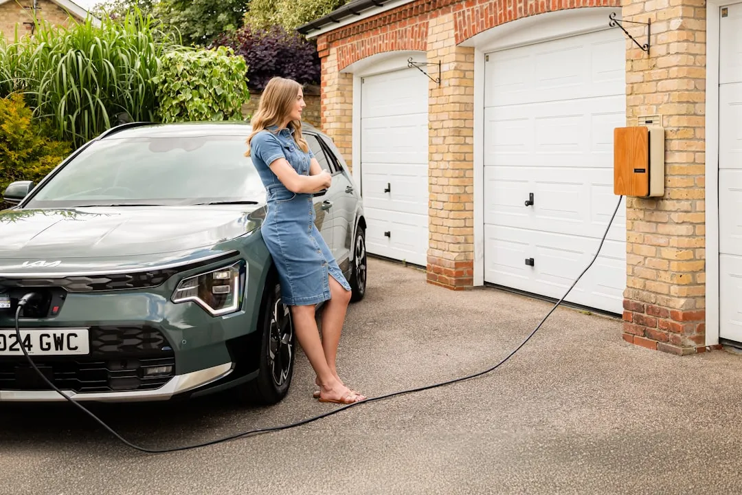 Electric vehicle plugged into orange wall-mounted charging unit outside a residential garage with person standing nearby