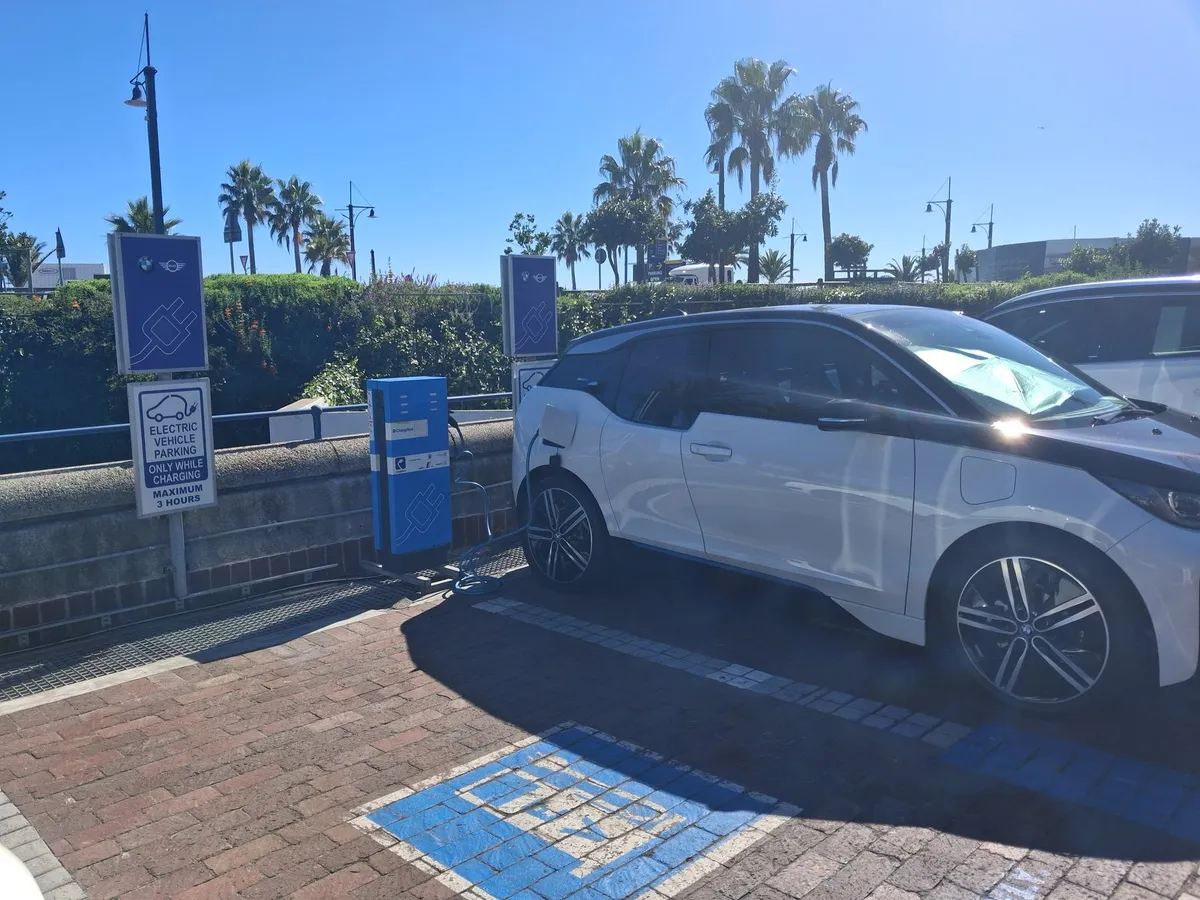 A white BMW i3 plugged into a public EV charger at the V&A Waterfront in Cape Town